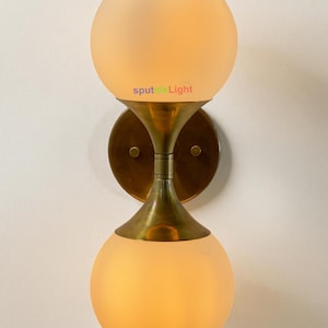 May include: A wall-mounted light fitting featuring two spherical, frosted glass shades. The fitting has a brass-coloured metal frame and a circular backplate. The text "sputnik Light" is visible.
