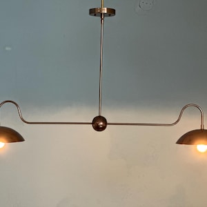 Puede incluir: Una lámpara colgante de dos luces de color bronce con un diseño minimalista. La lámpara presenta dos brazos curvos con pantallas en forma de cúpula, cada una con una luz cálida. La estructura central incluye una esfera y una varilla vertical para montaje en el techo.