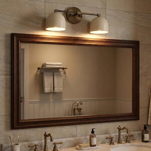 May include: A bathroom mirror with a dark brown wooden frame. Above the mirror is a two-light fixture with off-white shades and a brass finish. The mirror reflects a towel rack with towels and a sink area.