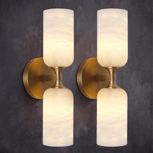 May include: A pair of wall sconces featuring a brushed brass finish and two cylindrical, white marbled glass shades each. The fixtures are mounted on circular brass backplates, with a central arm connecting the two vertical light sources.
