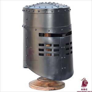 Puede incluir: Un yelmo medieval de caballero hecho de metal oscuro, remachado, con una visera con ranuras rectangulares para los ojos. El yelmo está montado sobre un soporte de madera, mostrando artesanía y diseño histórico.