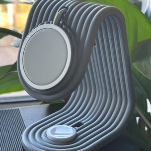 Peut inclure: Un support de chargement gris nervuré avec un socle de chargement circulaire. Le support a un design incurvé et maintient un appareil circulaire blanc. Un petit appareil circulaire blanc est à la base du support. Des feuilles vertes sont en arrière-plan.