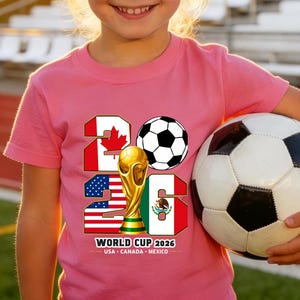 May include: A young person wearing a pink t-shirt with "2026" and a soccer ball graphic. The shirt also features the flags of the USA, Canada, and Mexico, and a gold trophy. The person is holding a soccer ball.