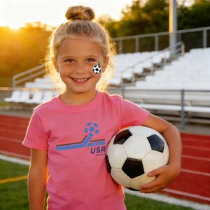 May include: A young person wearing a pink t-shirt with a soccer ball graphic and the text "USA". The person is holding a black and white soccer ball. The background includes a stadium and a sunset.