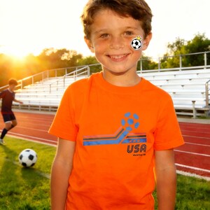 May include: An orange t-shirt with a soccer ball graphic and the text "USA World Cup 98". The shirt is worn by a young person on a soccer field. A soccer ball is on the grass.