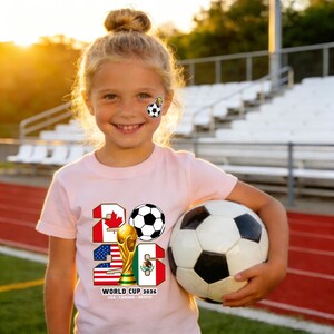 May include: A young girl wearing a light pink t-shirt with the text "2026" and a soccer ball graphic. The shirt also features the flags of Canada, the United States, and Mexico, along with a gold trophy. She is holding a soccer ball.