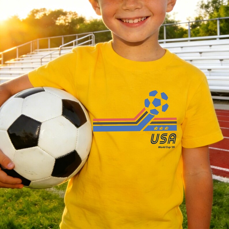 May include: A yellow t-shirt with a soccer ball graphic and the text "USA World Cup 26". The shirt is being held by a person and a soccer ball is in the person's other hand. The soccer ball is black and white.