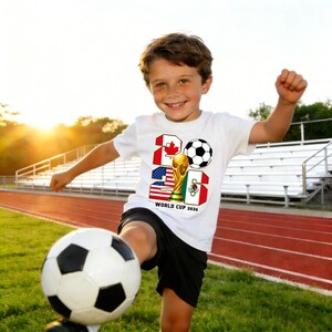 May include: A young person wearing a white t-shirt with a soccer ball graphic and the text "WORLD CUP 2026". The shirt features the flags of Canada, the United States, and Mexico. The person is kicking a soccer ball on a green field.