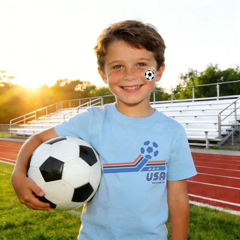 May include: A young person wearing a light blue t-shirt with a soccer ball graphic and the text "USA World Cup '20". The person is holding a black and white soccer ball. A soccer ball temporary tattoo is on the person's cheek.