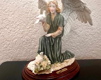 May include: A porcelain figurine of a kneeling angel with large white wings, wearing a dark green robe. The angel holds a white dove in one hand, while a second white dove rests on a cluster of cream-coloured roses. It sits on a dark wood base.
