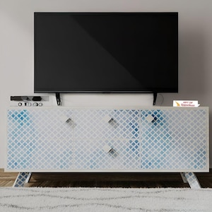 Puede incluir: Un mueble de TV blanco con un patrón geométrico azul y blanco. Tiene dos cajones y dos puertas con tiradores plateados, sostenidos por patas anguladas. Un televisor negro grande está montado encima. El mueble mide 150 cm de ancho.