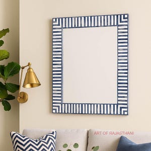 Puede incluir: Espejo de pared rectangular con marco a rayas azul marino y blanco. El espejo está colgado en una pared beige, con una lámpara de pared dorada a la izquierda. El espejo está sobre un sofá gris con cojines decorativos.