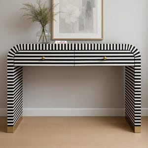May include: A modern console table with a black and white striped pattern. The table has two drawers with gold-colored knobs and gold accents on the legs. A framed artwork and a vase with greenery sit on top.