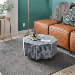 Peut inclure: Une table basse octogonale avec un motif géométrique bleu et blanc est posée sur un tapis gris. La table contient des objets décoratifs et une petite pancarte. Un canapé sectionnel en cuir marron est en arrière-plan.