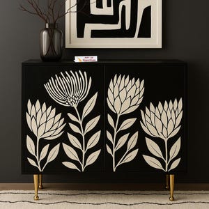 May include: Black cabinet with cream floral design on the doors. The cabinet has gold legs and a dark background. A framed black and white abstract art piece hangs above the cabinet. A vase with branches sits on top of the cabinet.