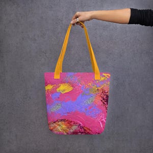 Może przedstawiać: R&oacute;żowa torba typu tote z ż&oacute;łtymi uchwytami, z abstrakcyjnym wzorem w odcieniach r&oacute;żu, fioletu, ż&oacute;łci i pomarańczy ze srebrnymi brokatowymi akcentami. Torba jest trzymana na szarym tle.