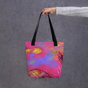 Może przedstawiać: R&oacute;żowa torba typu tote z czarnymi uchwytami. Torba ma abstrakcyjny wz&oacute;r z plamami ż&oacute;łtego, fioletowego i srebrnego brokatu. Torba jest trzymana przez osobę.