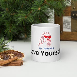 Könnte beinhalten: Weißer Keramikbecher mit dem Text "I'M POWERFUL Love Yourself" in Schwarz und Blau. Der Becher zeigt eine rote Umrissillustration einer Frau mit Sonnenbrille. Der Becher steht auf einer weißen Oberfläche.