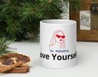 May include: White ceramic mug with the text "I'M POWERFUL Love Yourself" in black and blue. The mug features a red outline illustration of a woman wearing sunglasses. The mug is on a white surface.