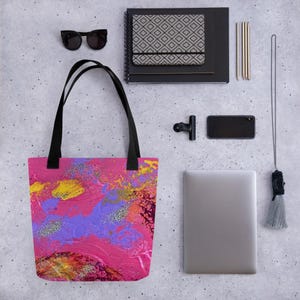 Może przedstawiać: R&oacute;żowa torba typu tote z czarnymi uchwytami, z abstrakcyjnym wzorem w kolorze ż&oacute;łtym, fioletowym i czerwonym ze srebrnymi akcentami brokatu. Torba jest prezentowana z okularami przeciwsłonecznymi, notatnikiem, telefonem i laptopem.