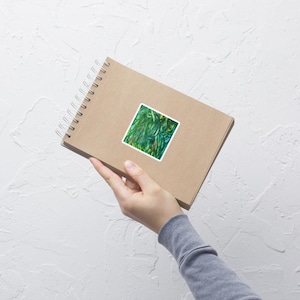 Può includere: Un adesivo quadrato con un design astratto nei toni del verde, blu e giallo. L'adesivo è attaccato a un taccuino marrone a spirale. Il taccuino è tenuto da una persona.