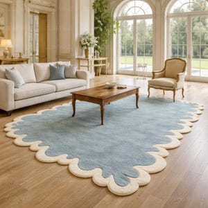 Könnte beinhalten: Hellblauer Teppich mit cremefarbenem, wellenförmigem Rand. Der Teppich befindet sich in einem Wohnzimmer mit Sofa, Couchtisch und Sessel. Das Design des Teppichs verleiht der Raumdekoration einen Hauch von Eleganz.