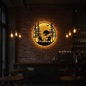 May include: A circular, black metal wall art piece showing a sunset scene with trees, chairs, and a bird. The art is illuminated with a warm yellow light, mounted on a brick wall. The scene suggests a relaxing outdoor theme.