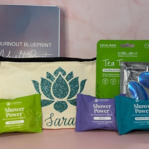 May include: A gift set featuring a spiral-bound notebook titled The Burnout Blueprint, a cream-colored zippered pouch with a teal glitter lotus design and the name Sara, a pink lip balm, a green facial mask, and three Shower Power aromatherapy tablets.