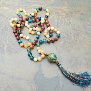 Puede incluir: Un collar de cuentas multicolores con un borla. Las cuentas son marrones, amarillas, azules y verdes. La borla es azul, dorada y marrón.
