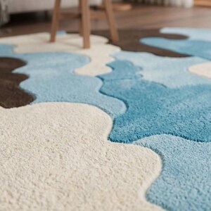 May include: A circular area rug featuring a tufted, high-pile texture with an abstract, organic pattern of interlocking shapes in shades of cream, light blue, and dark brown. The rug has a thick, plush surface with distinct color-blocked sections.