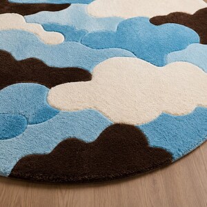 May include: A circular area rug featuring a tufted, cloud-like pattern in shades of dark brown, cream, and light blue. The design consists of overlapping, rounded shapes that create a textured, layered appearance across the entire round surface.