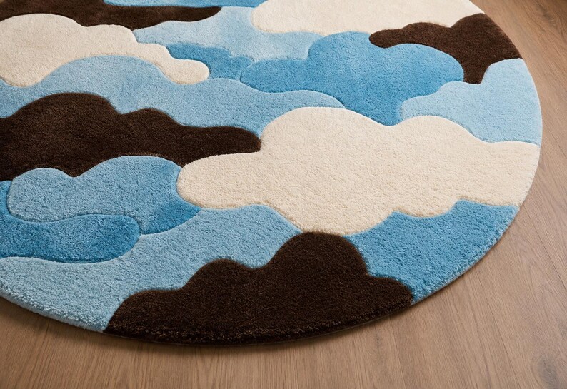 May include: A circular area rug featuring a tufted, cloud-like pattern in shades of dark brown, cream, and light blue. The design consists of overlapping, rounded shapes that create a textured, layered appearance across the entire round surface.