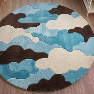 May include: A circular area rug featuring a layered, abstract cloud pattern in shades of light blue, medium blue, cream, and dark brown. The rug has a plush, textured surface with distinct, curved shapes that create a soft, multi-dimensional appearance.
