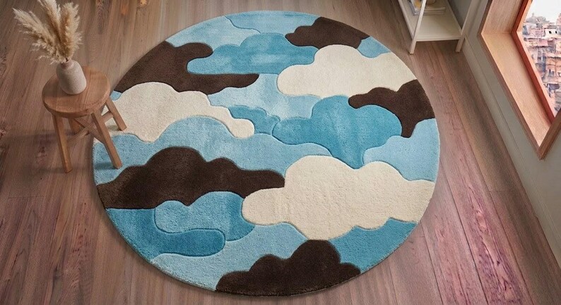 May include: A circular area rug featuring a layered, abstract pattern of clouds in shades of light blue, medium blue, cream, and dark brown. The design consists of soft, rounded shapes that overlap to create a textured, multi-colored surface.