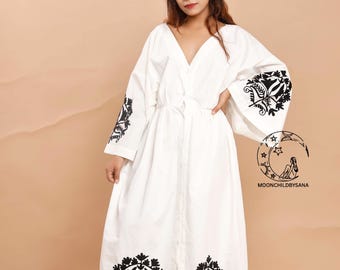 Op de afbeelding: Witte jurk met zwarte bloemenborduursels op de mouwen en zoom. De jurk heeft een V-hals en een strik in de taille. De mouwen zijn lang en uitlopend. De jurk is vloerlang en wordt gedragen met sandalen.