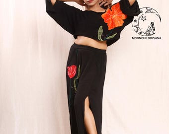 Può includere: Completo nero a due pezzi con ricami floreali. Il crop top presenta un grande motivo floreale arancione, e i pantaloni hanno uno spacco alto e un motivo floreale rosso. L'outfit è indossato con sandali. Il testo "MOONCHILDBYSANA" è visibile.