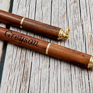 Puede incluir: Dos estuches de madera para puros con detalles de latón y llaveros. Uno de los estuches está grabado con el nombre "Greyson". Los estuches son de color marrón oscuro con una banda y herrajes dorados.