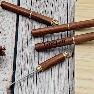 Puede incluir: Cuatro herramientas de cigarros de madera con detalles de latón se muestran sobre una superficie de madera. Una herramienta está grabada con el nombre "Greyson". Las herramientas son cilíndricas con una púa de metal que se extiende desde una. Una piña es visible en el fondo.