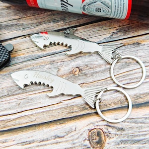 May include: Two silver shark-shaped bottle openers with key rings. One opener has the name "Haisley" engraved on it. The openers are on a wooden surface, with a beer bottle visible in the background.