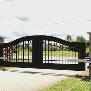 May include: A black wrought iron gate with a curved top and vertical bars. The gate is flanked by stone pillars and is set in a driveway with green grass and a cloudy sky in the background.