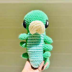 May include: A handmade crochet caterpillar plush toy. The caterpillar has a green head, a cream-coloured nose, and black and white eyes. The body transitions from light blue to green. The plush has multiple green legs.