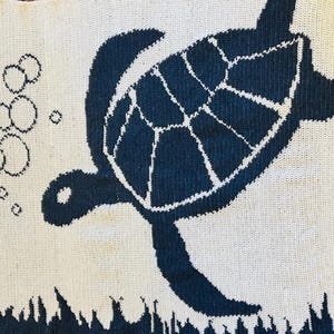 Könnte beinhalten: Gestrickter Stoff mit einem Design einer dunkelblauen Meeresschildkröte auf weißem Hintergrund. Die Schildkröte schwimmt, mit Blasen und Wasserpflanzen. Geeignet für die Inneneinrichtung.