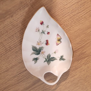 May include: White leaf-shaped serving dish with a gold rim and a handle. The dish is decorated with a strawberry plant design, including red strawberries, green leaves, and a yellow butterfly. A decorative piece for serving snacks or small items.