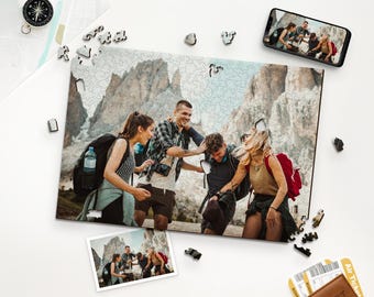 Op de afbeelding: Een houten legpuzzel met een foto van een groep mensen die in de bergen wandelen. De puzzel is gedeeltelijk in elkaar gezet, met enkele stukjes verspreid. Een smartphone toont dezelfde afbeelding. Andere items zijn een kompas, een foto en een portemonnee.
