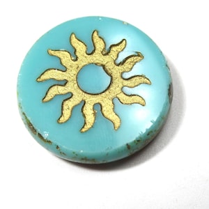 May include: A round, turquoise-colored bead with a gold sunburst design. The sun has a central circle and radiating wavy lines. The bead has a slightly textured surface and is set against a white background. This bead is suitable for jewelry making.