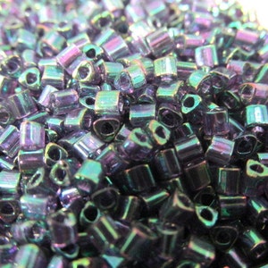 May include: A close-up of a large quantity of small, iridescent, dark purple and green glass beads. The beads are shaped like tiny, elongated hearts.