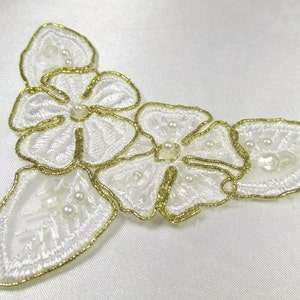 May include: Three white embroidered flowers with gold trim and pearl accents. The flowers are arranged in a triangular shape.