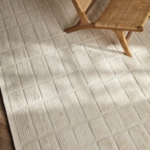 May include: A cream-colored area rug featuring a repeating grid pattern of raised rectangular blocks with vertical ribbed textures. The rug is partially covered by a wooden chair with a woven cane seat and backrest, showcasing a neutral palette.