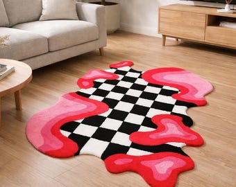 May include: A unique, abstract rug with a black and white checkered pattern at its center. The rug is bordered by a vibrant, layered design in shades of pink and red, creating a visually striking, modern aesthetic. The rug is placed on a wooden floor.
