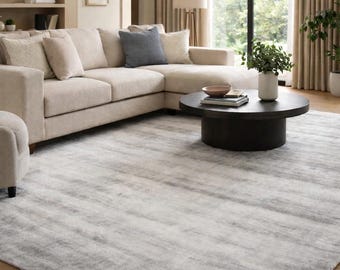 May include: A large, rectangular area rug with a soft, textured appearance. The rug features a blend of gray and white tones, creating a subtle, abstract pattern. The rug is placed in a living room setting, under a sectional sofa and a round coffee table.